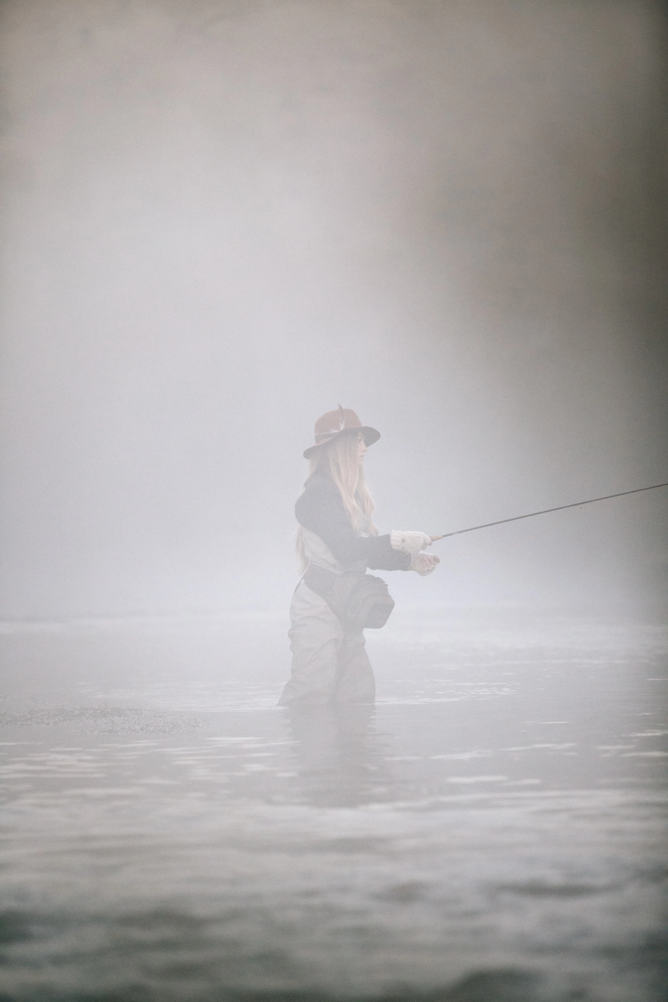A woman fly fishing in a foggy river, wearing a hat and waders, holding a fly rod.