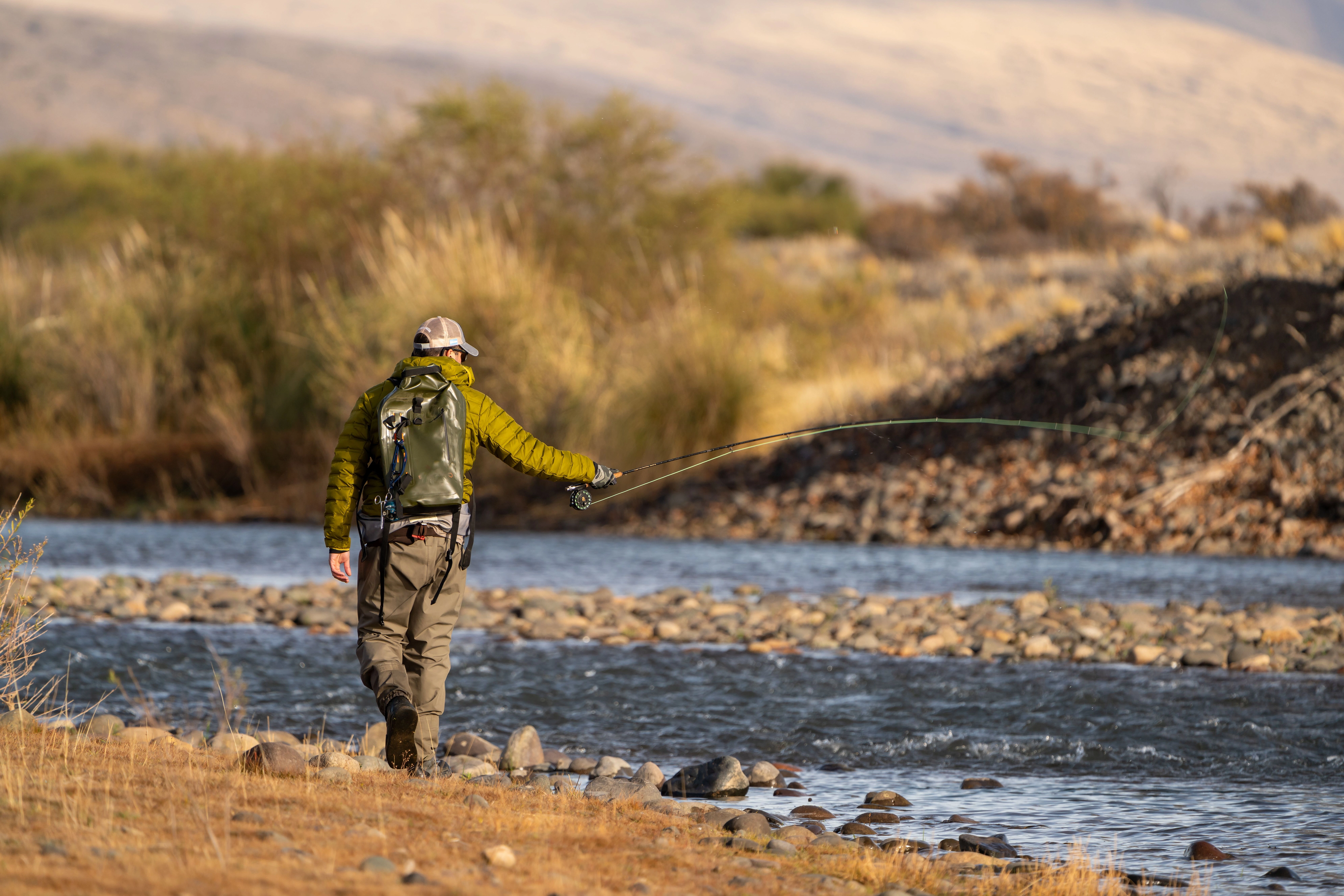 A fly fisherman walks along the bank of a rocky river with a slight ripple.