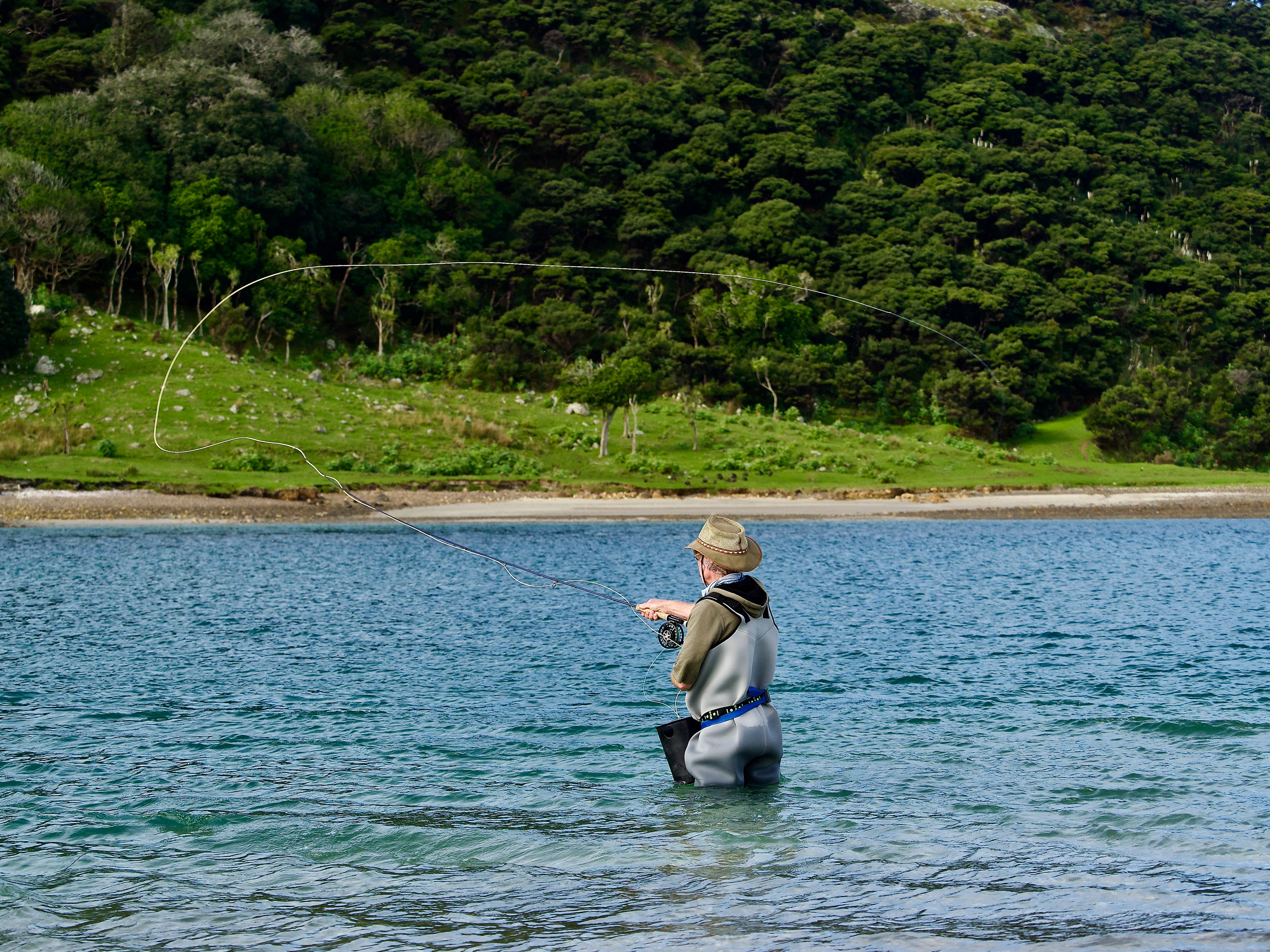 A fly fisherman wading in clear water, casting his line. A lush green hillside fills the background.
