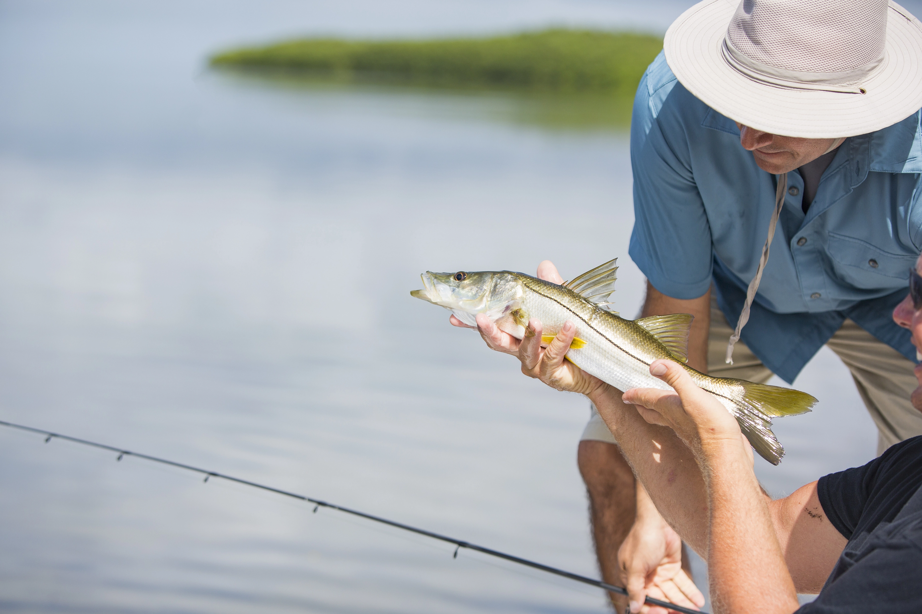 Close-up of two men holding a snook over calm water with a fishing rod in the foreground.